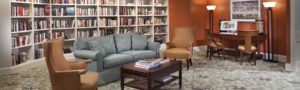 Brightly lit library with a wall of books and comfortable sofa and wing chairs